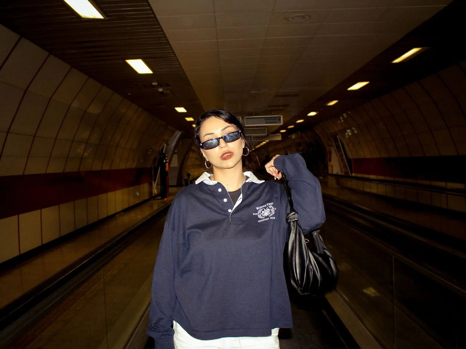 A fashionable woman wearing sunglasses and a navy shirt poses in an underground subway tunnel.